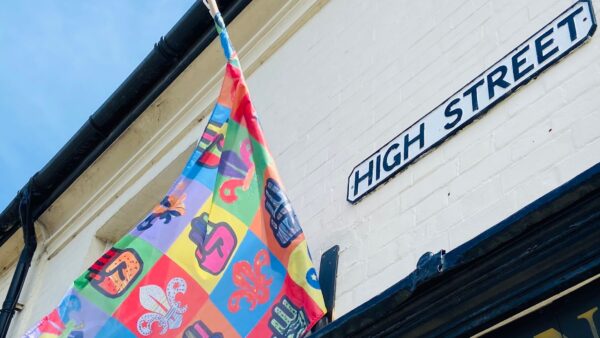 A colourful flag flying on a flagpole mounted onto a building with a High Street sign next to it