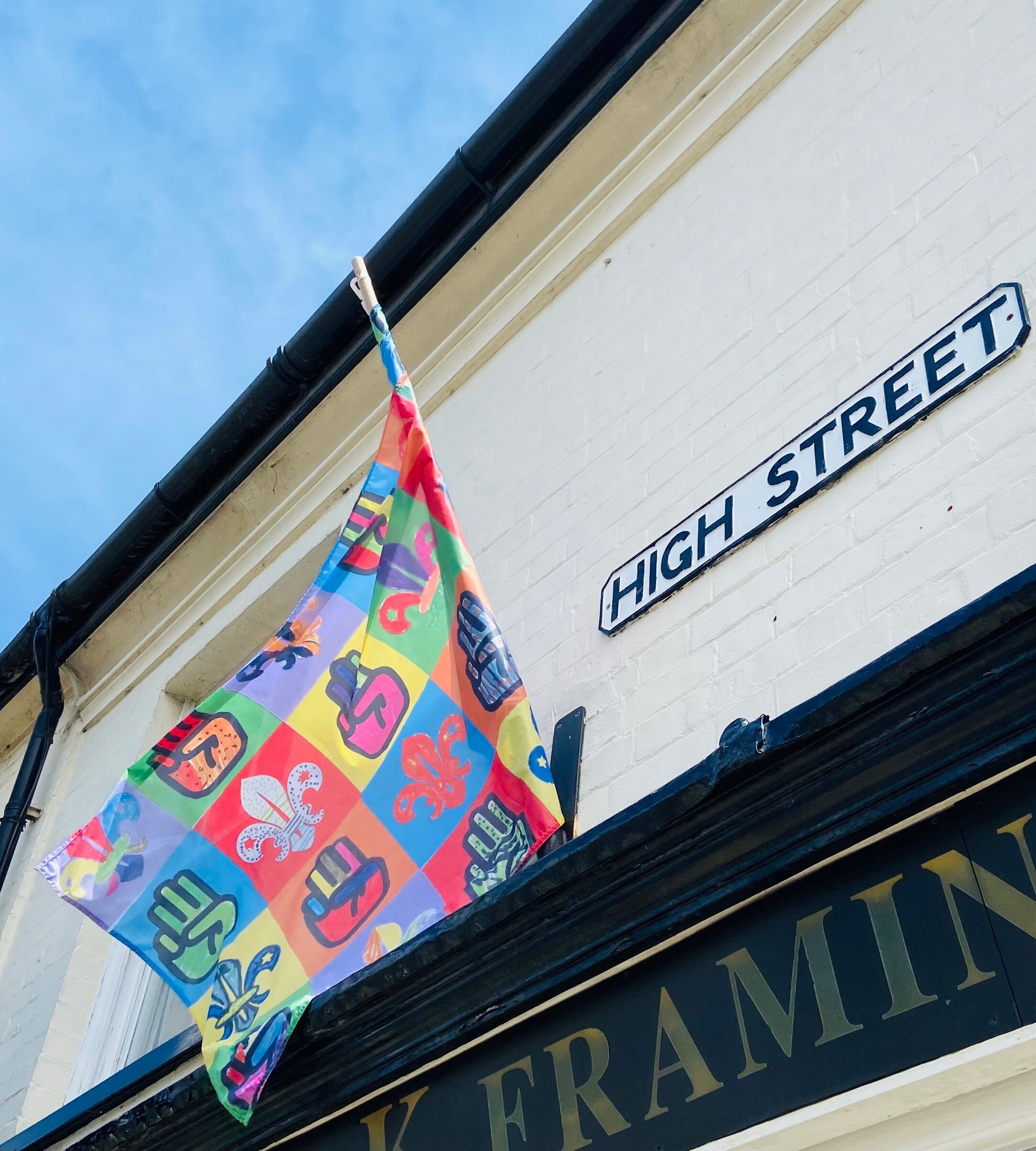 A colourful flag flying on a flagpole mounted onto a building with a High Street sign next to it