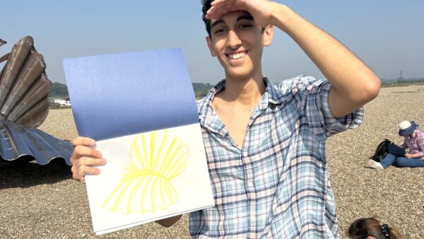 Boy standing on the beach showing his yellow drawing of a shell sculpture to the camera.