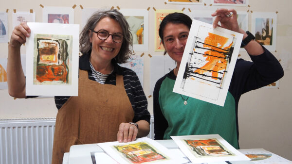 Two smiling women holding their vibrant orange, black and white risograph prints which have been scanned from their sketchbooks.