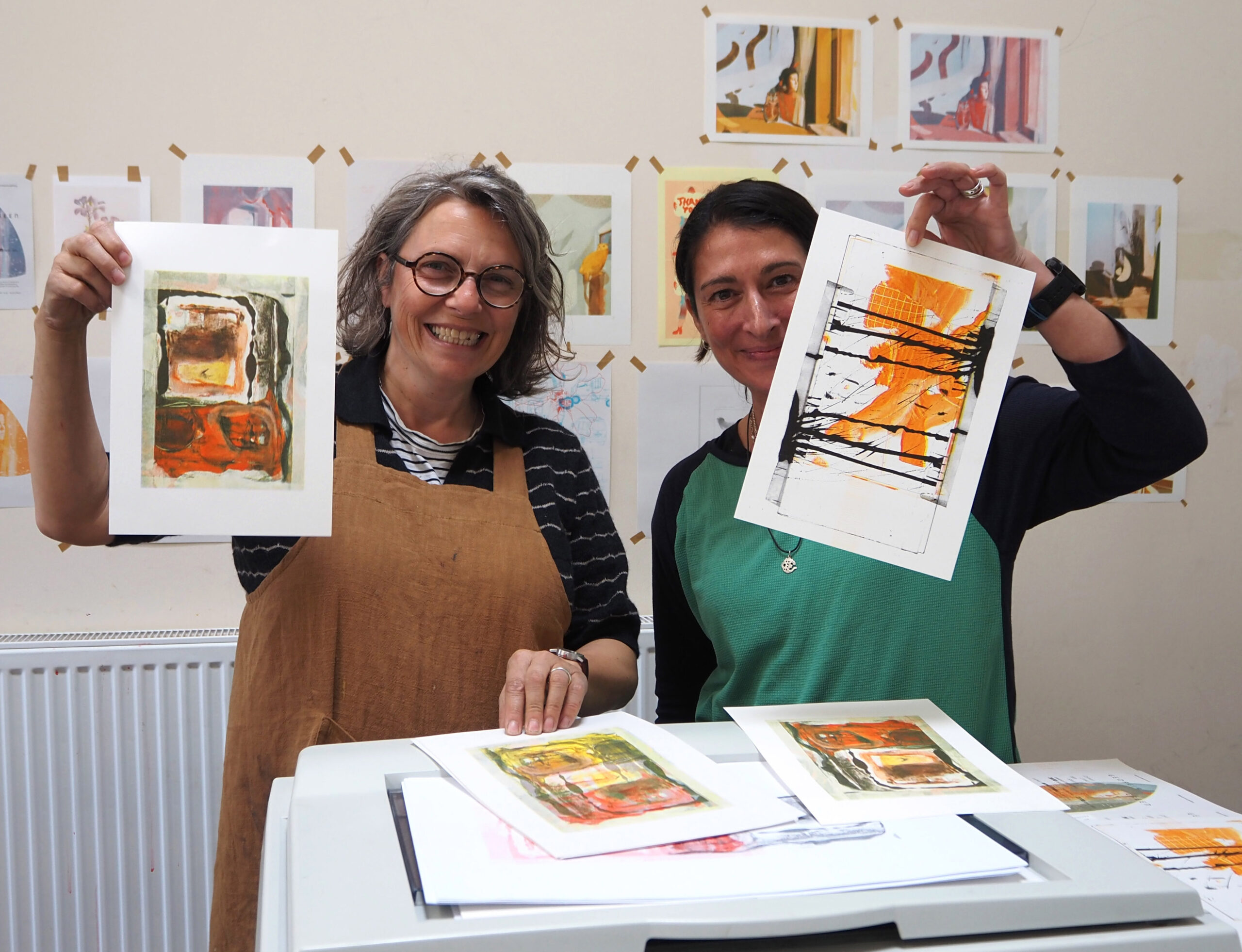 Two smiling women holding their vibrant orange, black and white risograph prints which have been scanned from their sketchbooks.