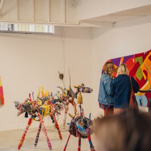 Photo of people looking around an exhibition, including a Birdseye view of a grand piano made out of red, yellow, purple and green felt, and goats sculpted from found materials like cans, bike wheels, and wool.